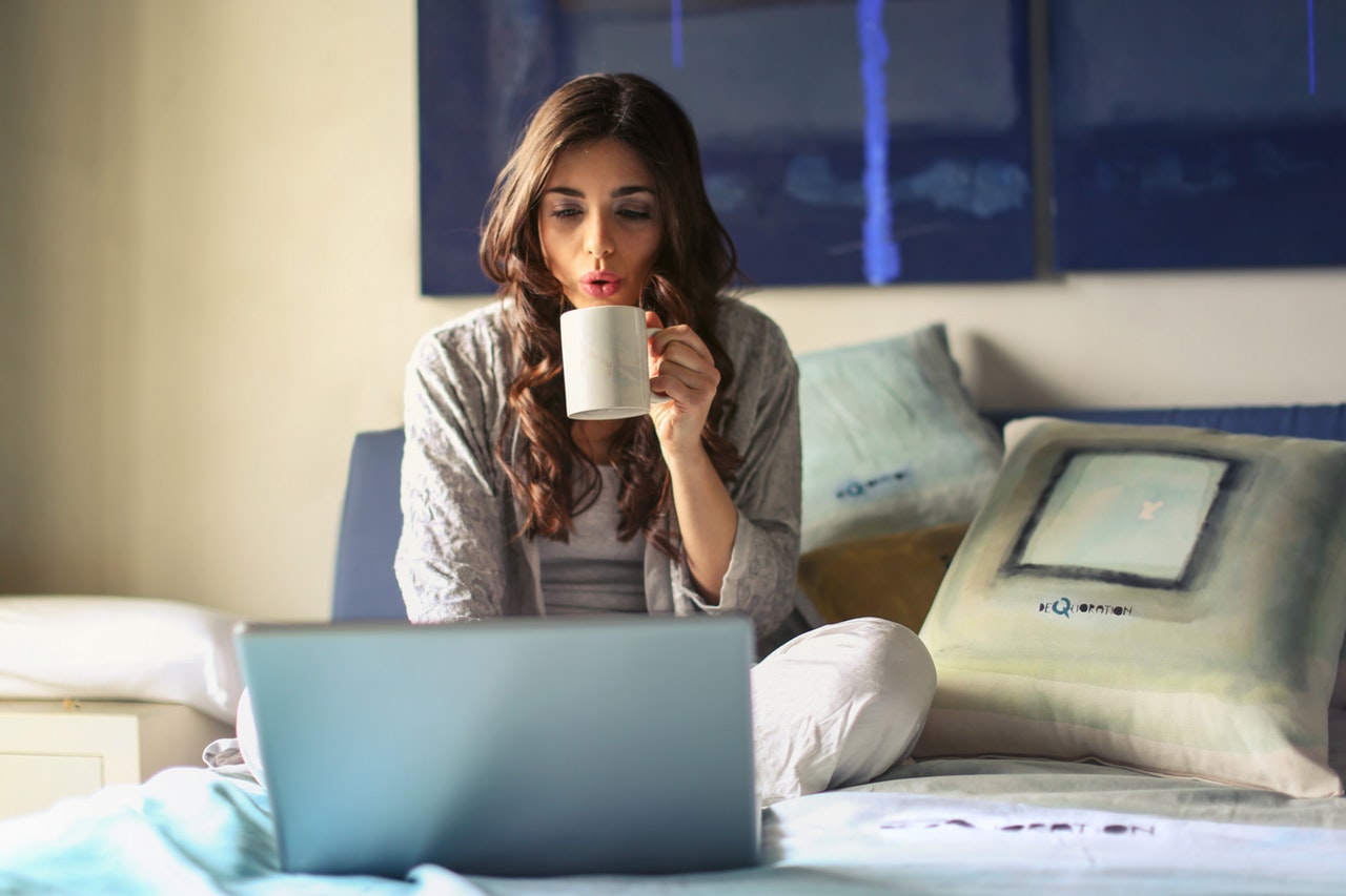 Woman sitting on bed with laptop holding mug to her mouth. She could be shopping online at Canada Pharmacy Depot!